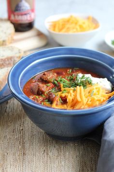 a blue bowl filled with chili and cheese next to bread on top of a table