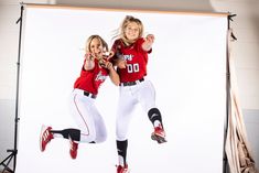 two girls in red and white baseball uniforms jumping up into the air