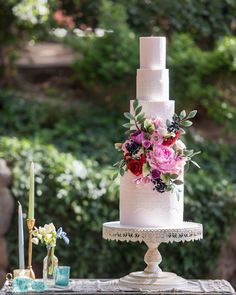 a white wedding cake with flowers on top sitting on a table in front of some bushes