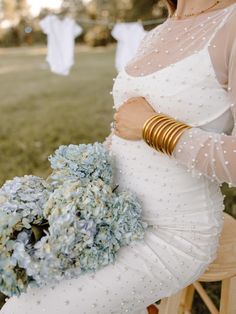 a woman in a white dress holding a bouquet of blue hydrangeas and gold bracelets