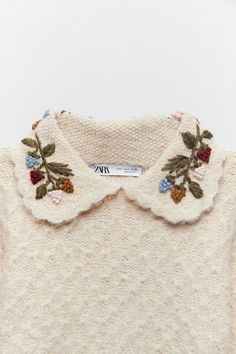 a white sweater with flowers and leaves on the collar is hanging from a hanger