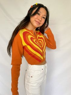 cropped orange cardigan with red, orange, and yellow heart design available in sizes xs, s, m, l model is 5'3" and wearing a size small Fitted Heart Print Sweater For Fall, Orange Fitted Long Sleeve Cardigan, Fitted Orange Long Sleeve Cardigan, Fitted Long Sleeve Orange Cardigan, Trendy Orange Crop Top For Fall, Casual Orange Crop Top For Fall, Orange Retro Long Sleeve Sweater, Retro Orange Long Sleeve Sweater, Orange Long Sleeve Retro Sweater