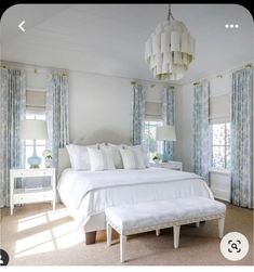 a white bed sitting in a bedroom under a chandelier next to two windows