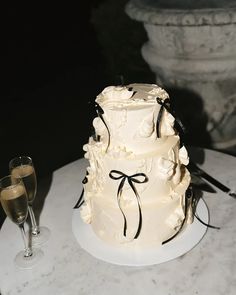 a three tiered wedding cake on a table with two champagne glasses and a statue in the background