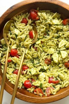 a wooden bowl filled with green pasta and cherry tomatoes next to two gold spoons