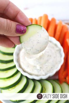 a hand holding a cucumber over a bowl of dip surrounded by sliced carrots