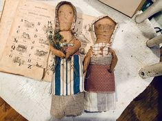 two wooden dolls sitting on top of a table next to an old book and plant