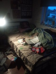 an unmade bed sitting next to a window in a room with lots of clutter on the floor