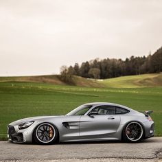 a silver mercedes sports car parked in front of a green field with hills and trees behind it