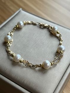 This delicate bracelet showcases a duo of 14k gold-plated chains, gracefully intertwined to form a double-layered embrace around the wrist. Adorned with six lustrous freshwater pearls, each sized at 7-8mm, the bracelet exudes a serene elegance. The pearls are evenly spaced, creating a harmonious balance with the shimmering gold links. This piece merges the timeless appeal of pearls with the modern finesse of layered chains, making it a versatile accessory for both casual and formal attire.