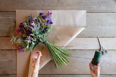 two hands are holding flowers and thread on a piece of brown paper with scissors next to it