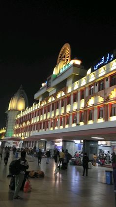 many people are walking around in front of a large building with lights on it at night