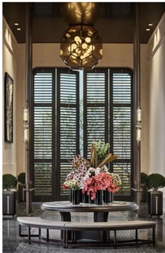 a table with some flowers on it in front of two large windows that have shutters