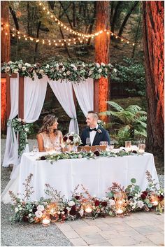 a bride and groom sitting at a table with candles in front of them, surrounded by greenery