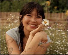 a woman sitting in a field with a flower on her arm and smiling at the camera