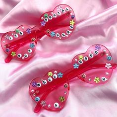 two pink heart shaped sunglasses with embellishments and flowers on the sides, sitting on a pink satin surface