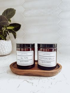 two jars of candles sitting on top of a wooden tray next to a potted plant