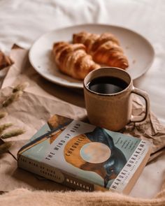 a cup of coffee next to a book and croissants on a bed