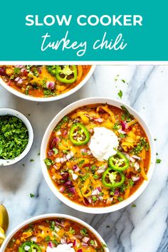 three bowls of slow cooker turkey chili on a marble table with spoons and cilantro