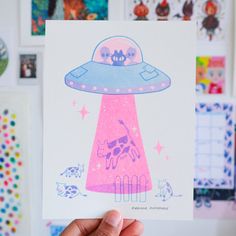 a hand holding up a pink and blue card with an alien ship in the middle