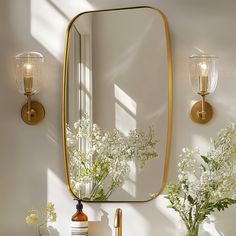 a bathroom sink with a mirror and flowers on the counter next to it, in front of a wall light