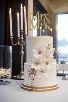 a white wedding cake with gold lettering and flowers on top is sitting on a table