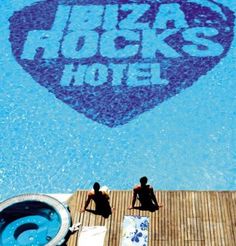 two people sitting on the edge of a swimming pool in front of a heart shaped sign