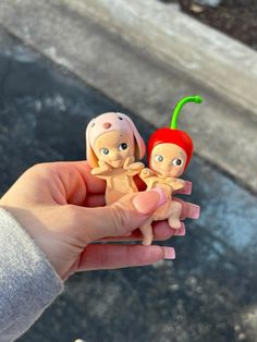 a person holding two small dolls in their hand, one with a cherry on it