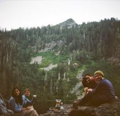 three people are sitting on the rocks with drinks in their hands and mountains behind them