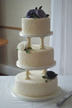 three tiered wedding cake with purple flowers on top and white frosting, sitting on a table