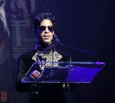 the man is speaking at an event wearing sunglasses and a black suit with jewels on his chest