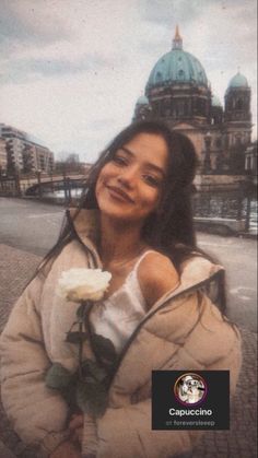 a woman holding a white rose in her right hand and looking at the camera with a building in the background