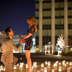 a man kneeling down next to a woman in front of many lit candles with buildings in the background