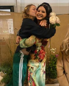 two women are hugging each other in front of a wall with flowers and greenery