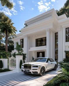 a white rolls royce parked in front of a large house with palm trees and bushes