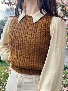 a woman standing in front of a tree wearing a brown sweater and white pants with her hands on her hips