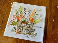 a close up of a card with carrots and a bunny in a basket on a wooden table