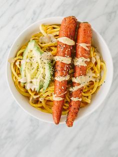 two carrots on top of noodles in a white bowl with sauce and avocado