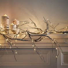 some candles are lit up on a mantel with branches in the foreground and lights strung across the mantle