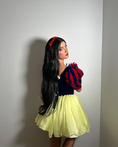 a woman with long black hair wearing a yellow skirt and red striped shirt standing in front of a white wall