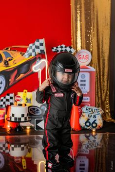 a young boy wearing a racing suit and helmet standing in front of a race car themed backdrop