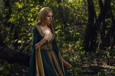 a woman in a green and gold dress is walking through the woods with her hand on her hip