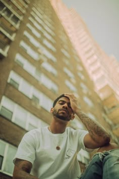 a man sitting in front of a tall building with his hand on his head while looking up at the sky