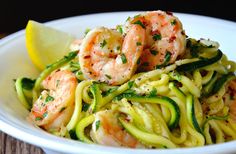 a white bowl filled with shrimp and zucchini noodles next to a lemon wedge