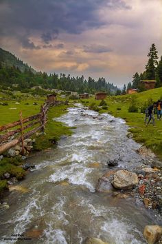 two people are standing on the edge of a river near a wooden fence and some rocks