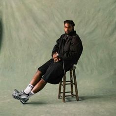 a man sitting on top of a wooden stool in front of a green wall with a clock