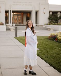 a woman standing on the sidewalk in front of a building with grass and flowers around her