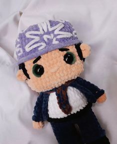 a crocheted stuffed animal wearing a purple hat and blue pants on a white sheet