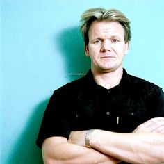 a man standing with his arms crossed in front of a blue wall and wearing a black shirt