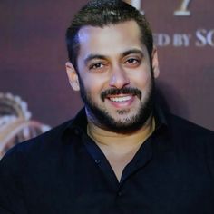 a man with a beard and black shirt smiling in front of a red carpeted wall